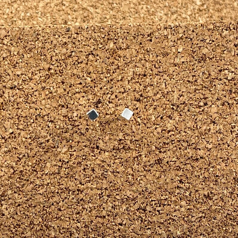 May include: Two small square earrings, one silver and one black, on a brown cork surface.