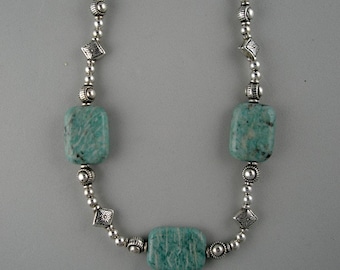 Op de afbeelding: Een zilveren ketting met groene edelsteen kralen. De ketting heeft vierkante kralen met een getextureerd oppervlak en ronde zilveren kralen.