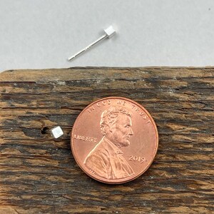 May include: A pair of small, silver, square stud earrings. The earrings are shown next to a US penny for scale.