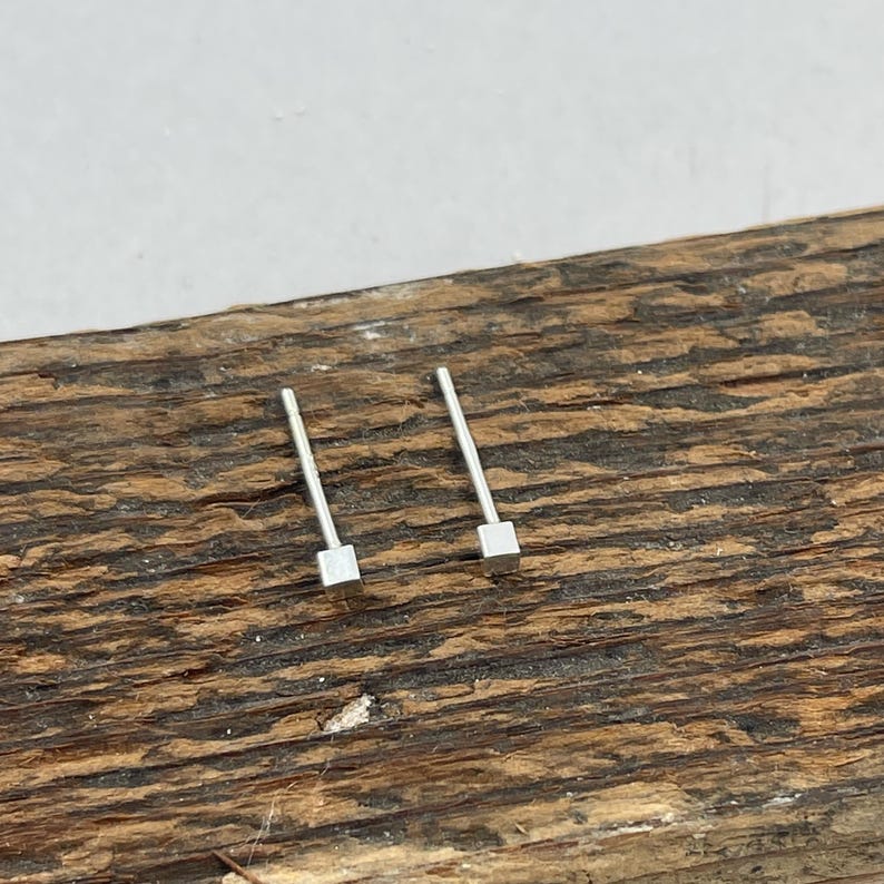 May include: A pair of silver square stud earrings on a wooden surface.
