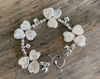 May include: A silver bracelet with a heart-shaped clasp and multiple white pearl heart charms.