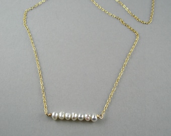 May include: A delicate gold necklace featuring a row of seven white pearls. The pearls are strung horizontally, suspended from a fine gold chain. The necklace is displayed against a neutral gray background.