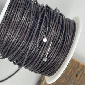 May include: A spool of dark brown waxed cotton cord. The cord is wound tightly around the spool, which is white with a black center. The cord is used for jewelry making, crafts, and other projects.