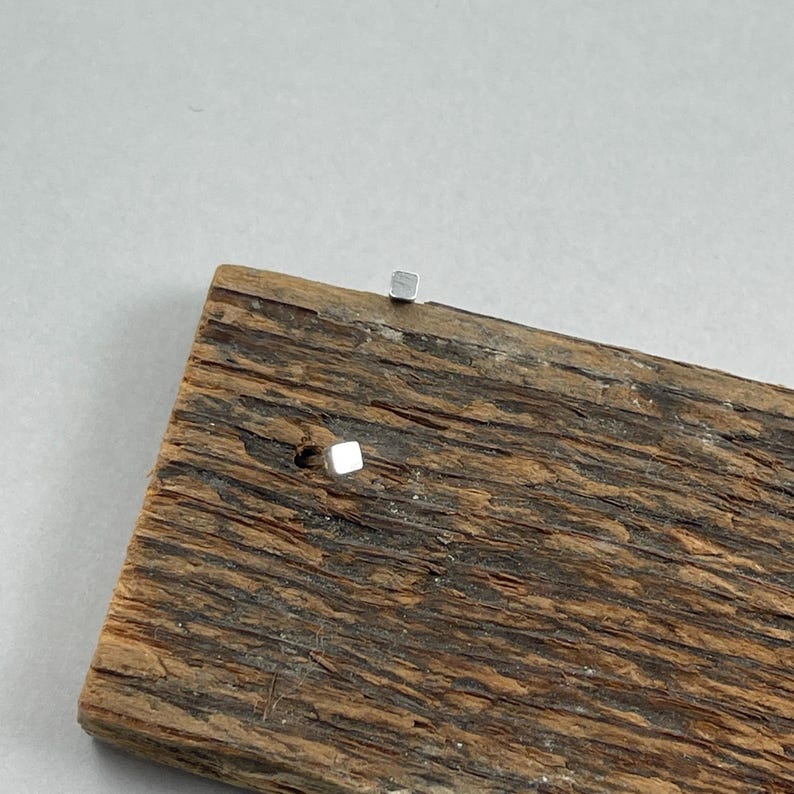 May include: A pair of small, silver square stud earrings on a piece of weathered wood.