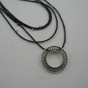 May include: A layered black beaded necklace with a silver circular pendant. The pendant has a delicate, intricate design.