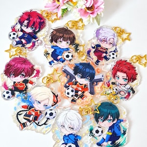Może przedstawiać: Kolekcja dziewięciu akrylowych breloków przedstawiających postacie z anime w stylu chibi w strojach piłkarskich, każdy ze złotym zapięciem w kształcie gwiazdy. Postacie są ukazane w dynamicznych pozach z piłkami nożnymi.