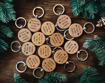 May include: A collection of wooden keyrings with silver hoops, each engraved with the German phrase "Mit dir im Team ist alles möglich" and a star. The keyrings are displayed on a dark wooden surface, with pine branches.