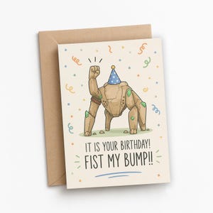 Può includere: Un biglietto d'auguri con una creatura rocciosa color marrone chiaro che indossa un cappellino da festa blu a pois e solleva il pugno. Il biglietto include coriandoli colorati e il testo: IT IS YOUR BIRTHDAY! FIST MY BUMP!! Con busta in carta kraft.