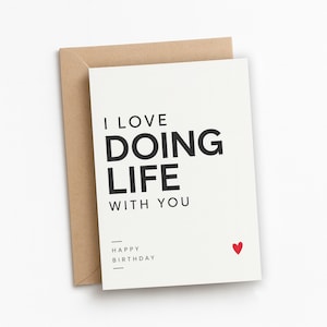 May include: A white greeting card with black text that reads I LOVE DOING LIFE WITH YOU and HAPPY BIRTHDAY with a small red heart icon. The card is placed in front of a plain brown envelope, creating a simple and clean visual presentation.