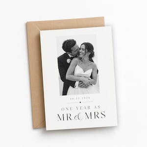 May include: A white greeting card with a black and white photograph of a couple embracing. The text reads 08.22.2026, ONE YEAR AS MR & MRS. The card is placed against a brown envelope. The design features clean typography and a small heart icon.