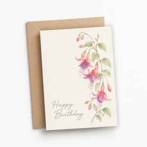 May include: A cream-colored greeting card with a watercolor illustration of fuchsia flowers in shades of pink and purple with green leaves. The text "Happy Birthday" is printed in a dark gray script font. A brown kraft paper envelope is behind it.