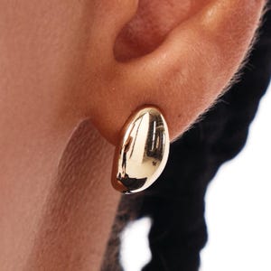 May include: A close-up of a gold-colored earring. The earring is a smooth, oval shape with a polished, reflective surface. The earring is positioned on an ear, with the focus on the jewelry.