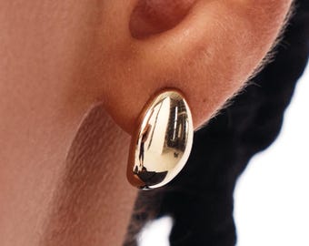 Op de afbeelding: Een close-up van een goudkleurige oorbel. De oorbel heeft een gladde, ovale vorm met een gepolijst, reflecterend oppervlak. De oorbel bevindt zich op een oor, met de focus op de sieraden.