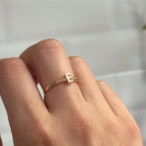 May include: A gold ring featuring a single letter 'E' set with small clear stones. The ring is worn on a finger.