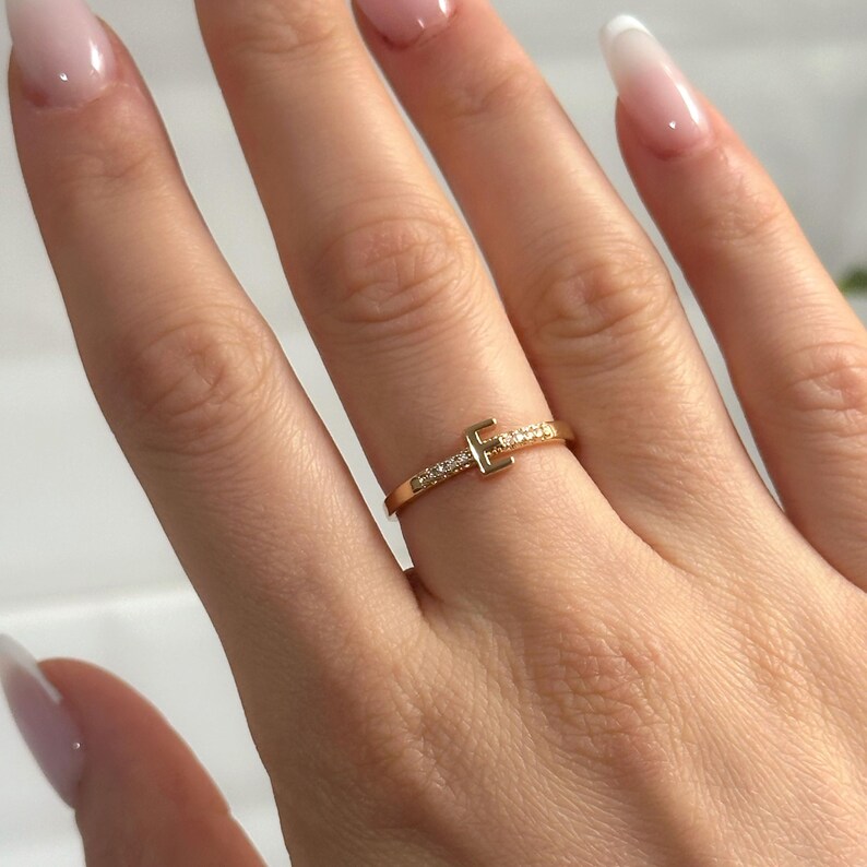 May include: A gold ring featuring a single letter 'E' set with small diamonds. The ring is worn on a finger.