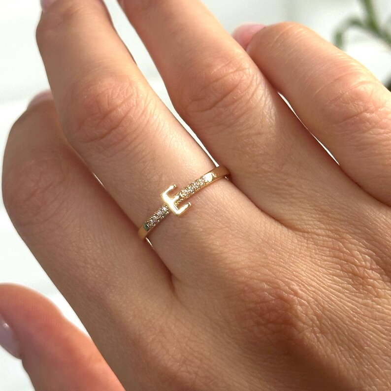 May include: A gold ring with a single letter 'E' set with small diamonds. The ring is on a finger.