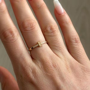 May include: A gold ring with a small letter "E" set with tiny clear stones. The ring is on a finger.