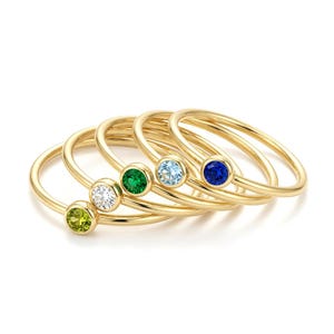 May include: A set of five thin gold rings, each featuring a single round bezel-set gemstone. The stones are colored in shades of lime green, clear white, emerald green, light blue, and deep royal blue, arranged in a cascading overlapping stack.