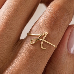 Op de afbeelding: Een gouden ring met een dunne, gepolijste band die een cursieve hoofdletter A vormt als centraal designelement. Het metaal heeft een gladde, reflecterende afwerking en de letter is direct geïntegreerd in de doorlopende ronde band.