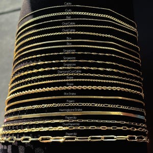 May include: A collection of gold-colored necklaces displayed against a dark background. The necklaces vary in style, including cable, curb, ball, oval cable, box, Singapore, diamond cut cable, rope, box snake, figaro, herringbone snake, and paperclip designs.