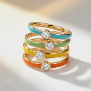 May include: A stack of four gold rings, each featuring a single round clear gemstone set in a bezel. The bands are finished with colorful enamel in shades of turquoise, light green, yellow, and orange, arranged in a vertical column formation.