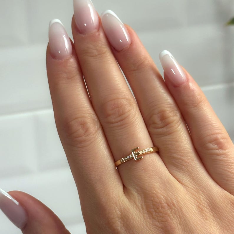 May include: A gold ring with a small diamond-encrusted letter 'T' on a finger with a white French manicure.