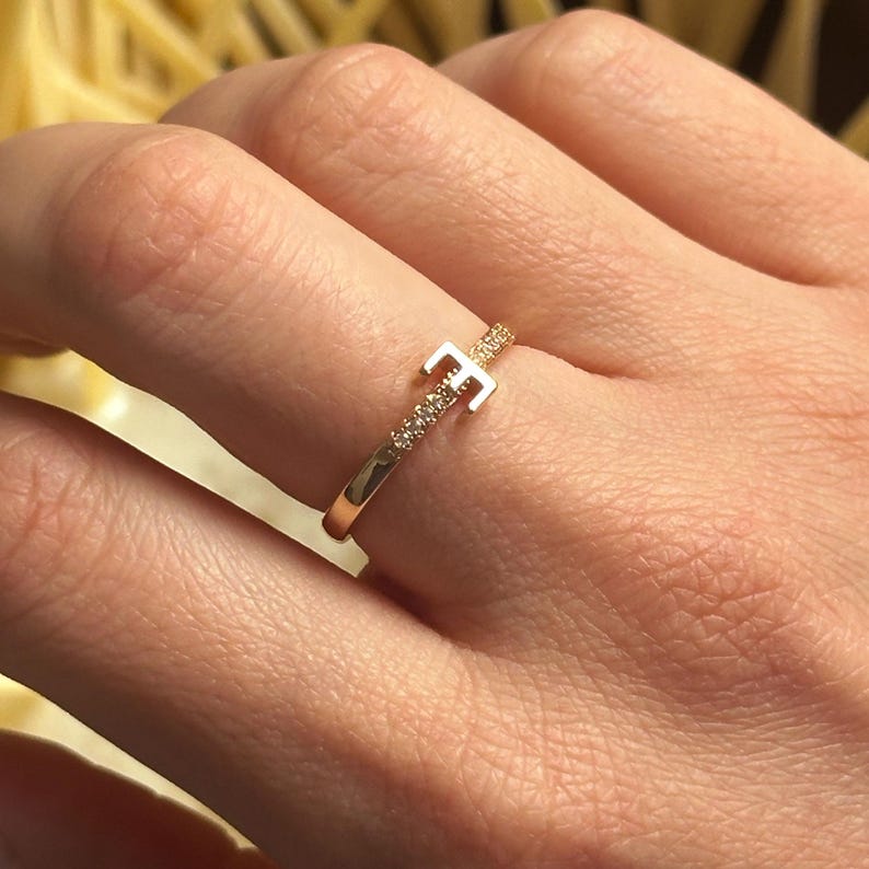 May include: A gold ring with a single letter 'E' set with small clear stones. The ring is on a finger.