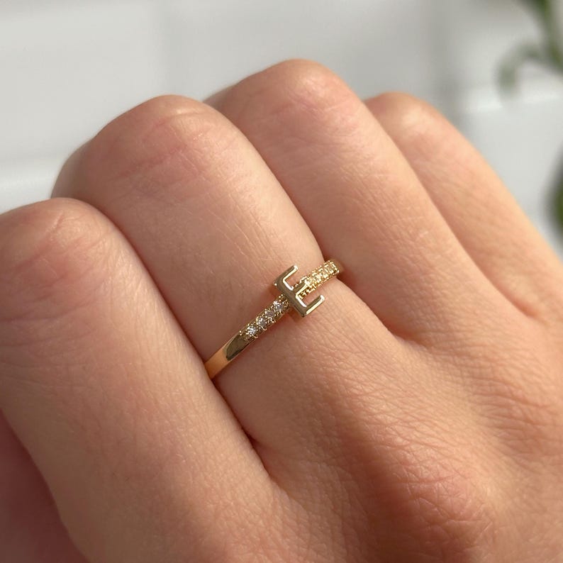 May include: A gold ring featuring a single initial 'E' set with small clear stones. The ring is worn on a finger.