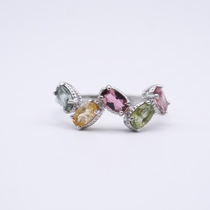 May include: A silver ring with a row of oval gemstones in shades of pink, yellow, green, and blue-green. The gemstones are set in a row, creating a colourful and elegant design. The ring is a piece of jewellery.