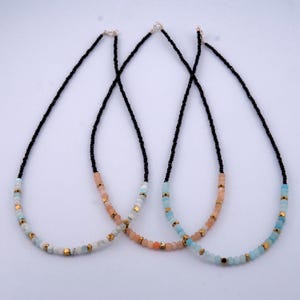 May include: Three delicate necklaces with black beads and colorful gemstone accents. The necklaces feature a gradient of light blue, peach, and gold beads.