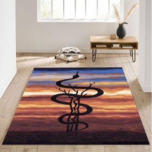 Op de afbeelding: Een rechthoekig vloerkleed met een zwart slangmotief dat zich rond een boom wikkelt tegen een zonsondergang. Het kleed heeft tinten blauw, oranje en bruin, wat een dramatisch effect creëert. Geschikt voor woon- en slaapkamers.