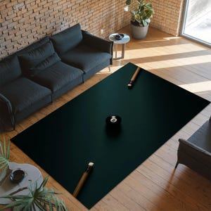 May include: A rectangular, dark green rug with a pool table design. The rug features an eight ball and two pool cues. The rug is placed on a hardwood floor in a living room setting. The room has a black sofa and a brick wall.