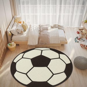 Op de afbeelding: Een ronde vloerkleed die lijkt op een voetbal. Het kleed heeft een zwarte achtergrond met witte vijfhoeken en zwarte lijnen, wat het klassieke voetbalpatroon creëert. Het ligt op een lichte houten vloer in een slaapkamer.