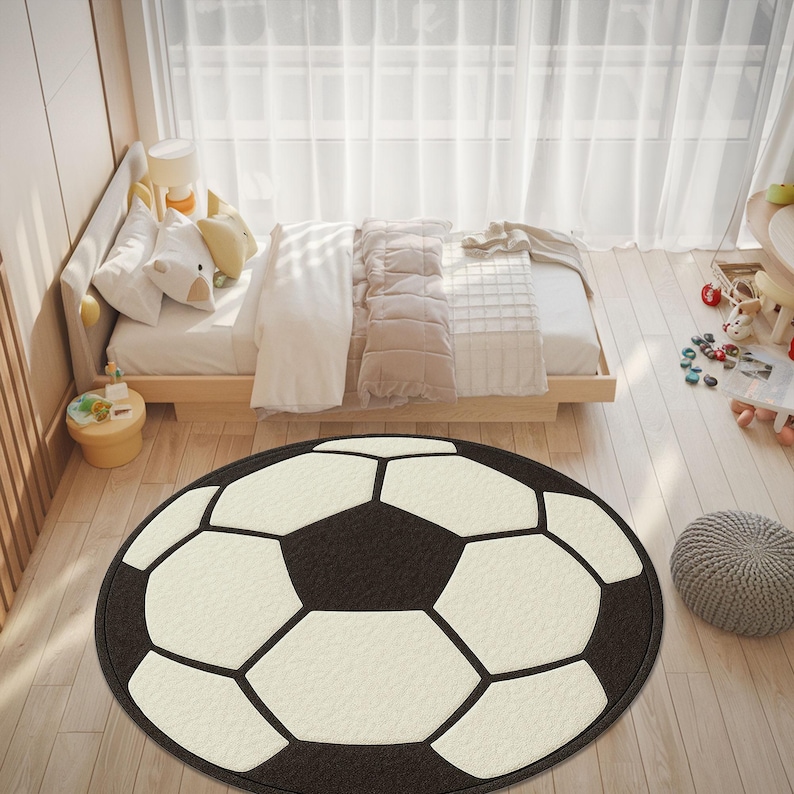 Puede incluir: Una alfombra redonda con dise&ntilde;o de bal&oacute;n de f&uacute;tbol. Presenta un fondo negro con pent&aacute;gonos blancos y l&iacute;neas negras, reproduciendo el patr&oacute;n cl&aacute;sico. Se encuentra sobre un suelo de madera clara en un dormitorio.
