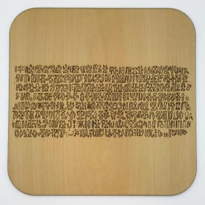 May include: A light brown wooden coaster with rounded corners. The coaster features a long, rectangular section of black, stylized text. The text appears to be a series of symbols and characters.