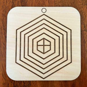 May include: A light-colored wooden coaster with a geometric design. The design features a series of concentric hexagons, with a cross shape at the center. A small hole is at the top for hanging.
