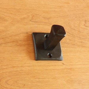 May include: Black metal hook with a rectangular base and a single hook. The hook is mounted on a wooden surface.