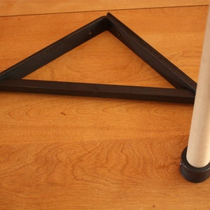 Op de afbeelding: Een zwarte metalen plankbeugel in de vorm van een driehoek met afgeronde randen. De beugel is bevestigd aan een houten paal met een zwarte kunststof voet.