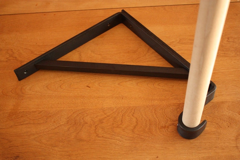 Op de afbeelding: Een zwarte metalen plankbeugel in de vorm van een driehoek met afgeronde randen. De beugel is bevestigd aan een houten paal met een zwarte kunststof voet.