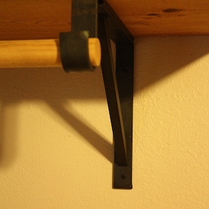 Op de afbeelding: Een houten plank met een zwarte metalen beugel. De plank is met schroeven aan de muur bevestigd. De beugel heeft een driehoekige vorm en is van metaal.
