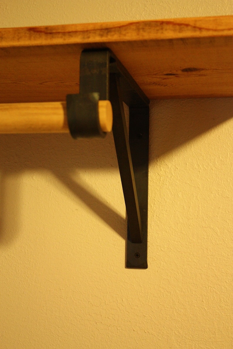 Op de afbeelding: Een houten plank met een zwarte metalen beugel. De plank is met schroeven aan de muur bevestigd. De beugel heeft een driehoekige vorm en is van metaal.
