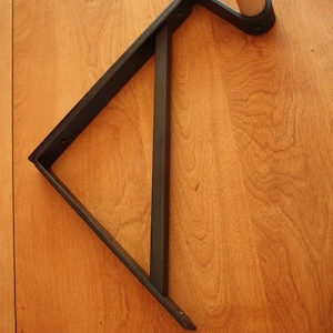 Op de afbeelding: Een zwarte metalen plankbeugel in de vorm van een driehoek. De beugel is bevestigd aan een houten oppervlak.