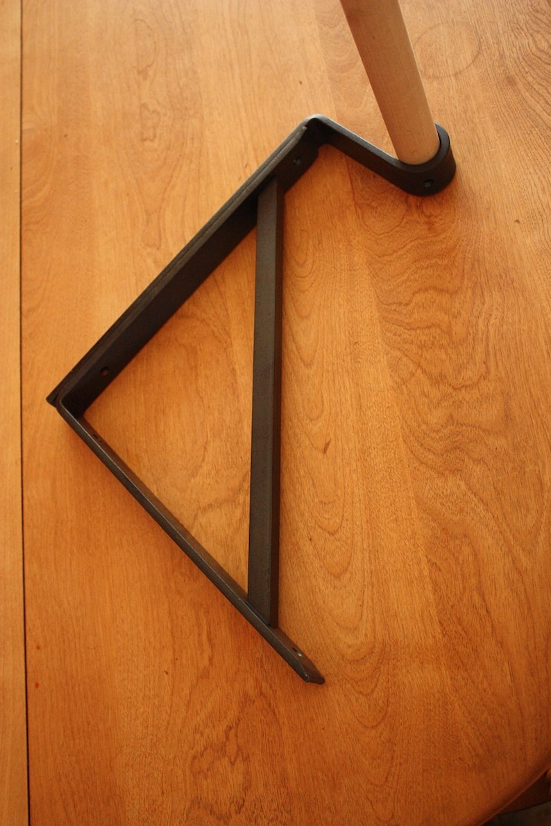 Op de afbeelding: Een zwarte metalen plankbeugel in de vorm van een driehoek. De beugel is bevestigd aan een houten oppervlak.