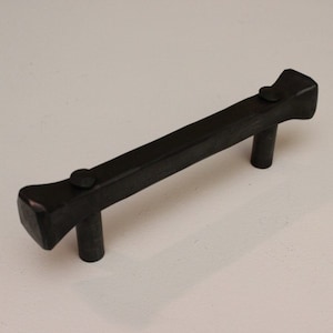 May include: Black metal cabinet pull with a squared-off design. The pull has a flat, rectangular shape with rounded edges and two small, round rivets.
