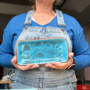 May include: A rectangular, turquoise ceramic tray with a floral design. The tray has a brown rim and is held by a person wearing blue overalls and a blue sweater. The floral design features detailed outlines of various flowers.