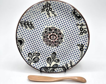 May include: A white ceramic plate with a blue polka dot pattern and black and red floral designs. A wooden butter knife is included.