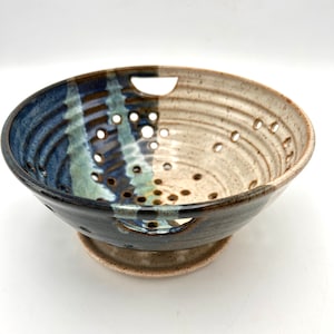 May include: A handmade ceramic colander with a two-tone design. The left side is dark blue with lighter blue and brown streaks, while the right side is tan. It has numerous small holes and two large handle cutouts.