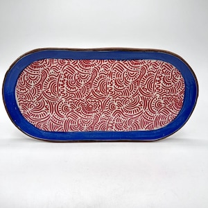 May include: Oval ceramic serving tray with a blue border and a red and white patterned interior. The tray has a brown rim and is set against a white background. The tray is a decorative piece for serving food or displaying items.