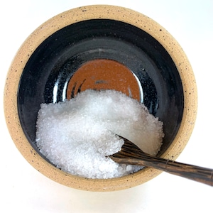 May include: A small, speckled brown ceramic bowl with a black and brown glaze inside. The bowl is filled with white salt crystals and a wooden spoon is partially inserted into the bowl.