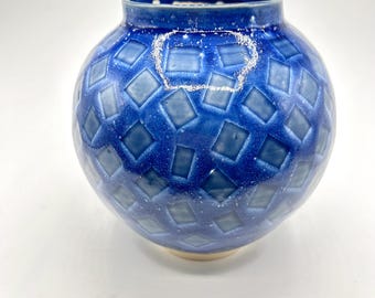 May include: A round, blue ceramic vase with a diamond pattern. The vase has a wide, rounded body and a narrow neck. The glaze is a deep, glossy blue with lighter blue accents. The base is a light beige color.